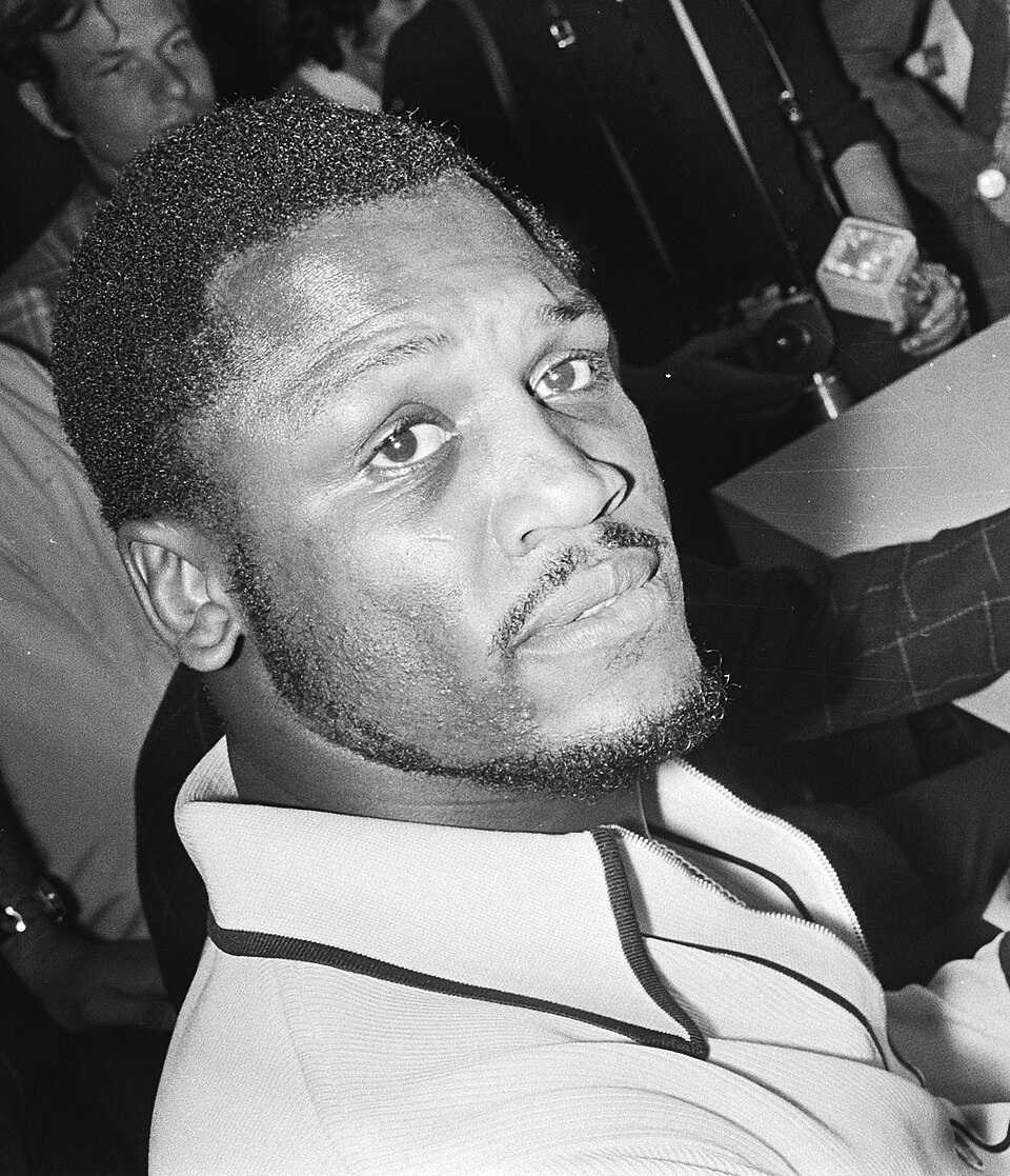 Joe Frazier reading Dutch newspaper about his singing career at Amsterdam Airport Schiphol (cropped)