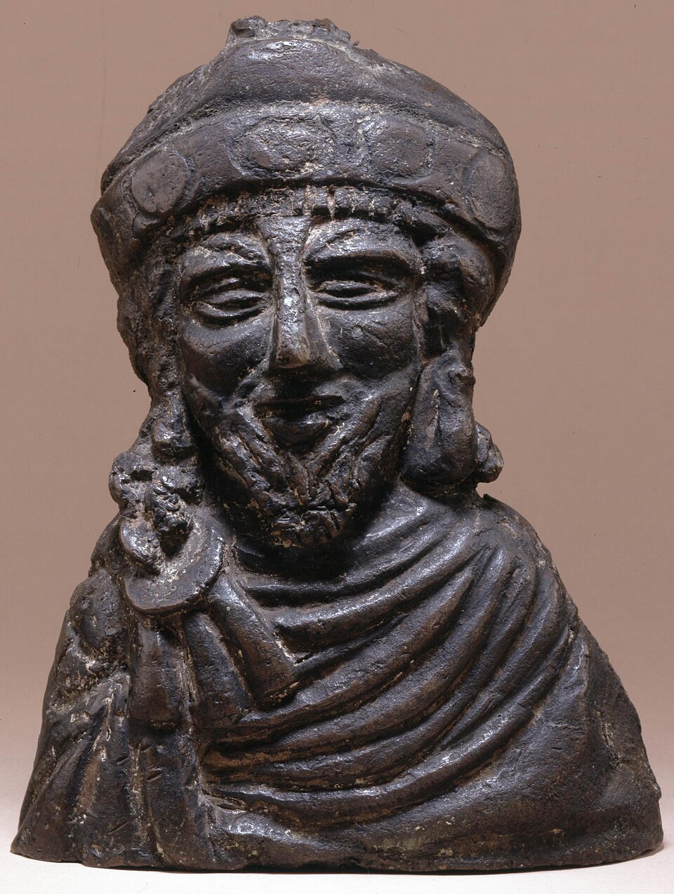 Steelyard weight; bronze, filled with lead; in the form of the bust of a bearded emperor (Phocas?) with long hair, wearing a diadem, and a chlamys fastened…